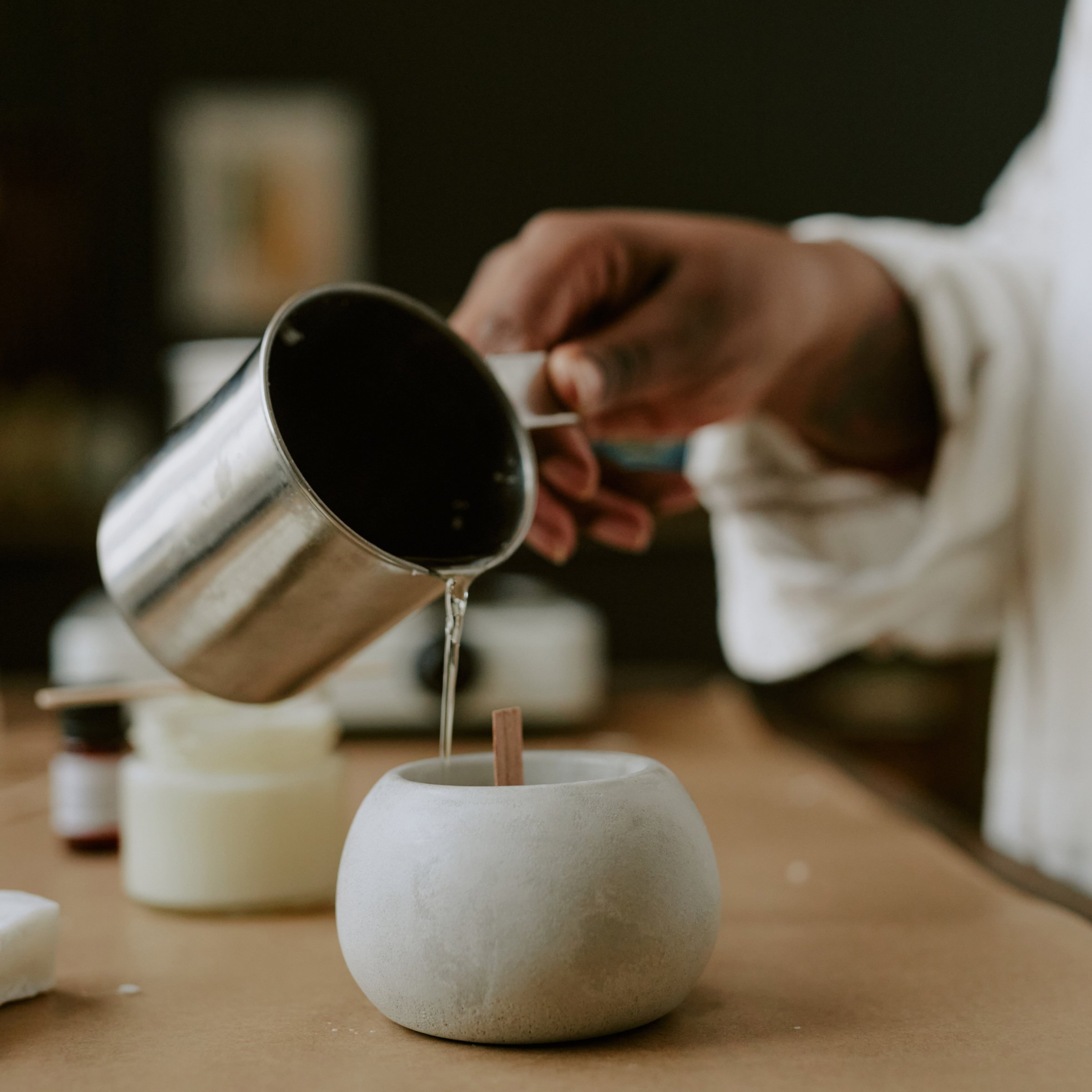  A close-up shot of a person pouring melted wax from a metal pitcher into a small, round container, likely to create a candle. 