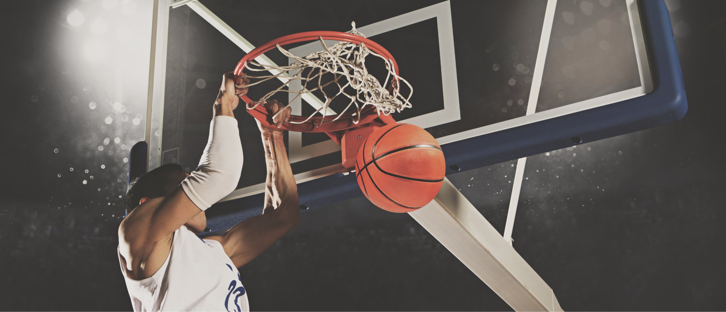 A basketball player is captured mid-air, performing a powerful dunk during a game. The player hangs onto the rim with one hand as the basketball is about to pass through the hoop. The action takes place under bright stadium lights, emphasizing the intensity and energy of the moment. The scene conveys athleticism, focus, and the excitement of basketball.