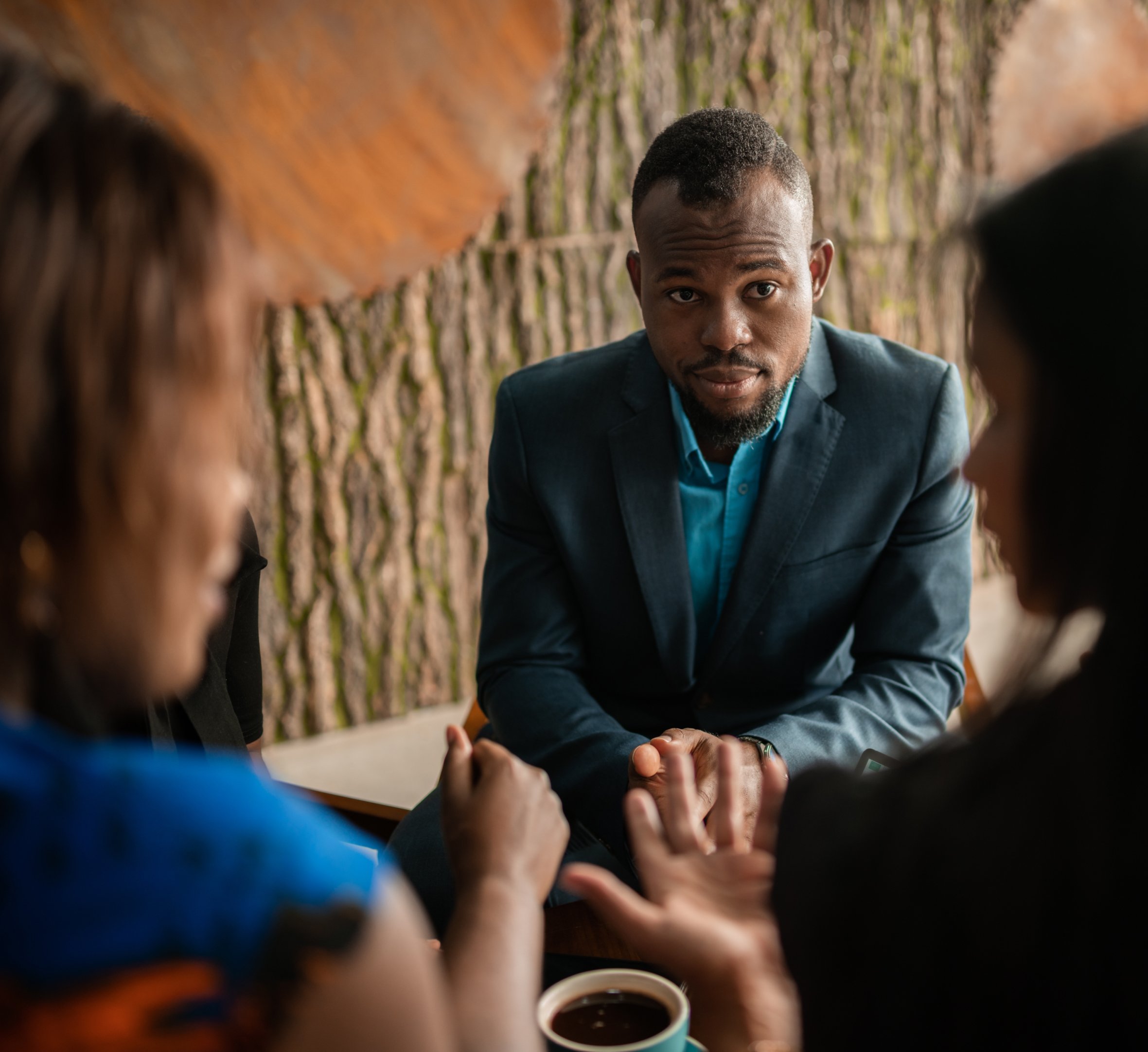 A man in a blue suit sits attentively during a discussion with two women, listening intently as one gestures. The setting has a natural, earthy backdrop, creating a warm and professional atmosphere.