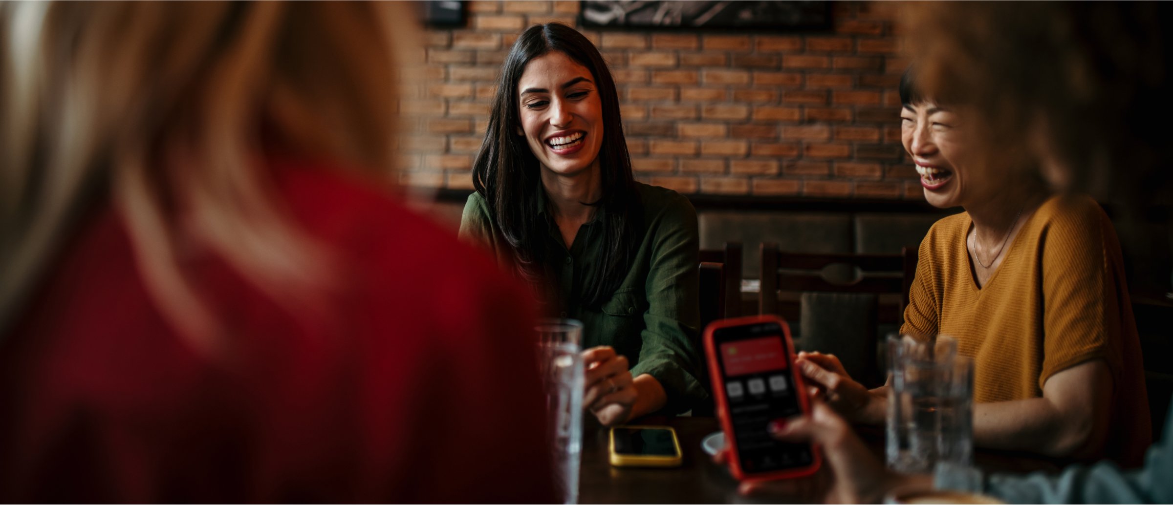 A group of friends share a moment of laughter while sitting at a table in a cozy restaurant. A woman in a green shirt smiles, with others engaged in the conversation, enjoying their time together.