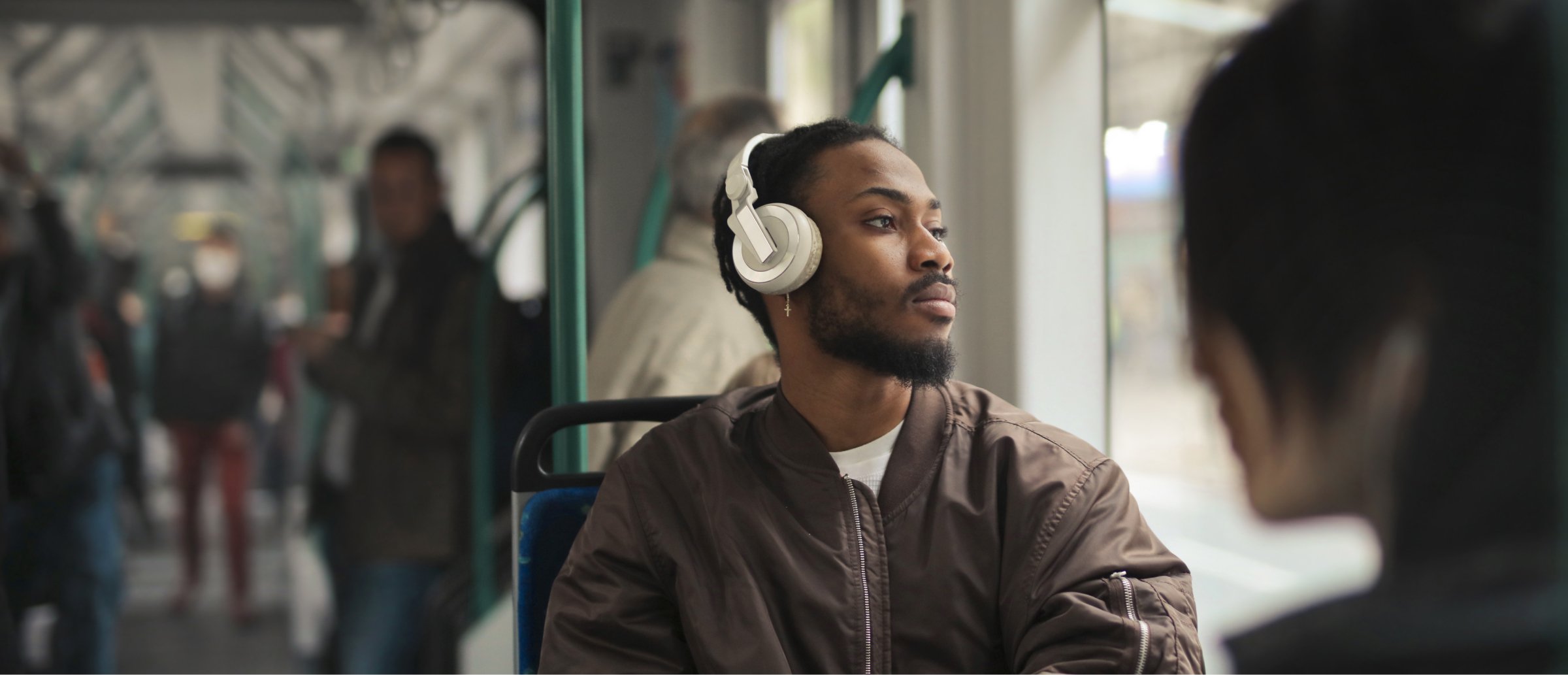 A young man with a beard is sitting on public transportation, wearing large white headphones and looking thoughtfully out the window. He is dressed casually in a brown jacket, and the soft light from outside highlights his profile. In the background, other passengers are visible, some standing and others seated, creating a typical busy public transit atmosphere. The image conveys a sense of calm and reflection during the commute, with the man enjoying music or a podcast through his headphones.