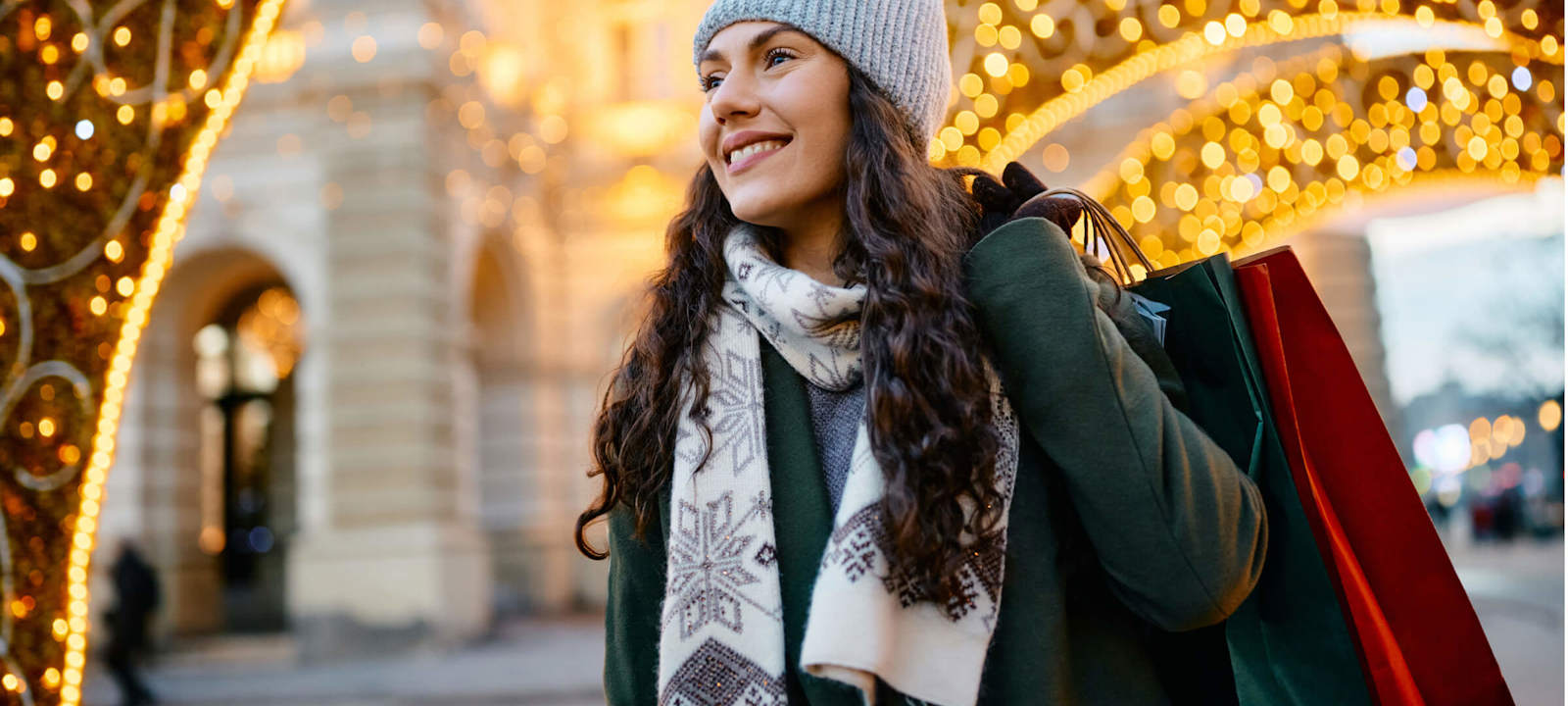 A smiling woman dressed in winter clothing, holding shopping bags while walking under festive holiday lights. She is wearing a knit hat, scarf, and coat, enjoying a seasonal shopping experience in a brightly lit, decorated urban setting.