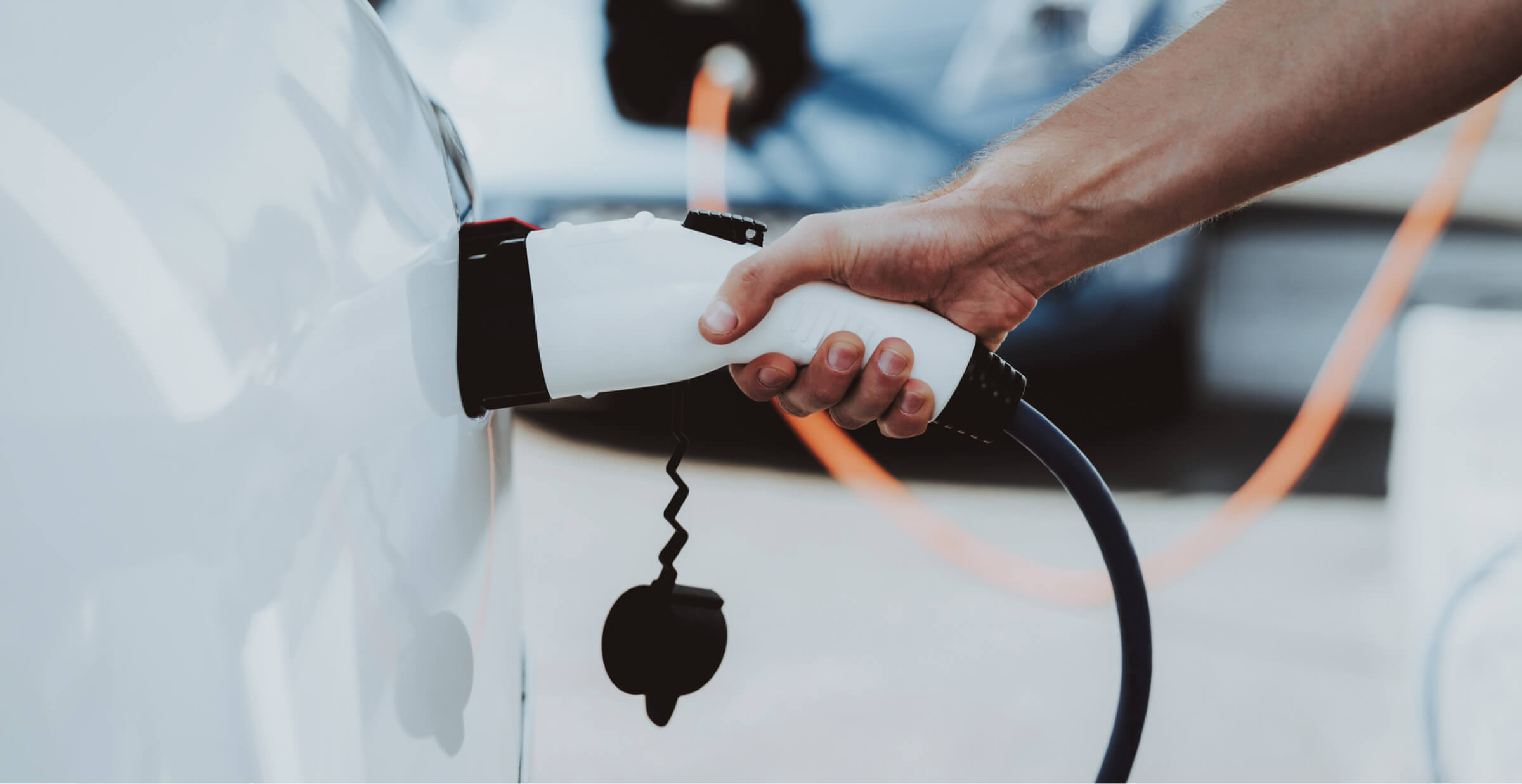  A close-up of a person’s hand plugging a charging cable into an electric vehicle. The white car is connected to a charger through a thick blue cable, indicating the process of recharging the car’s battery. In the background, another electric vehicle is visible with its own charging cable. The scene highlights the growing adoption of electric vehicle technology.