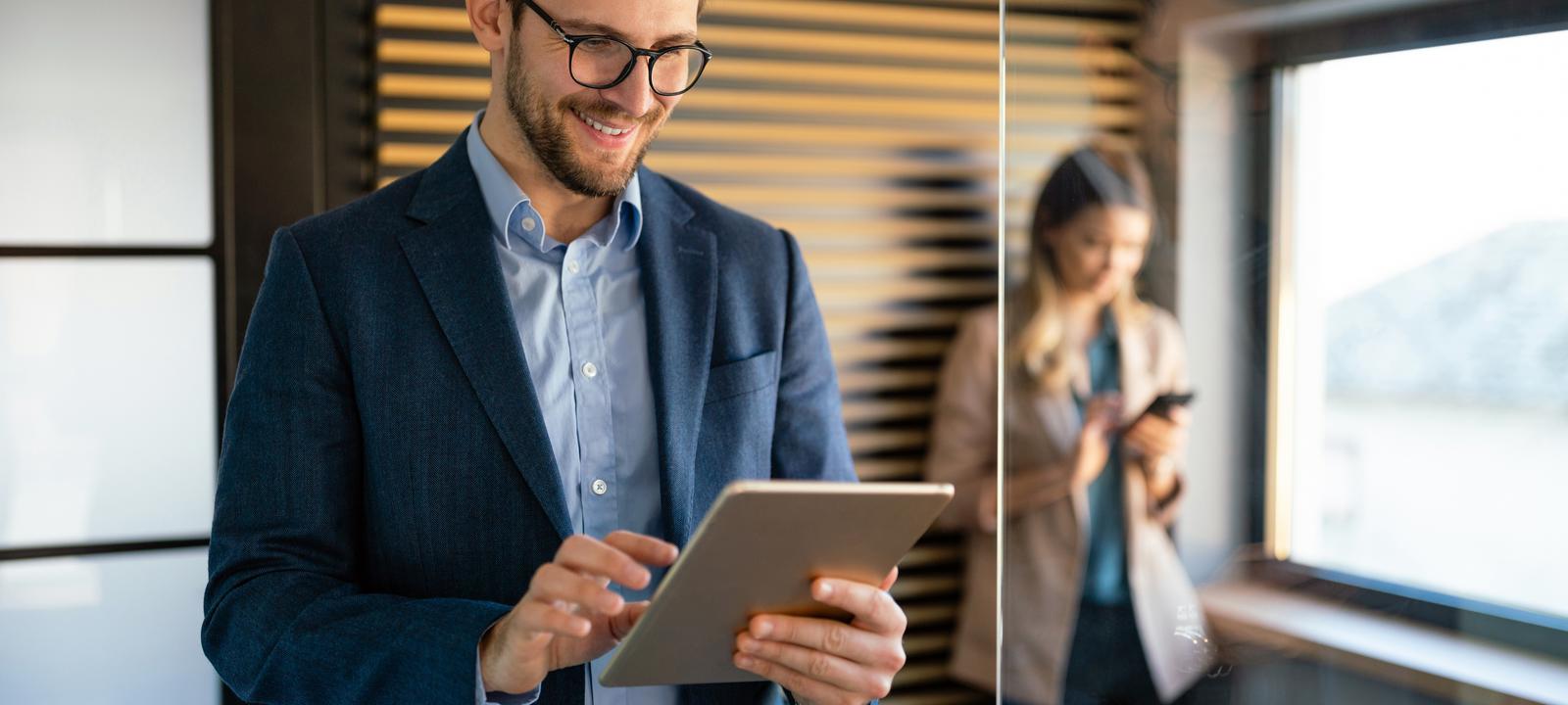 A man in a business suit and glasses is smiling while working on a tablet in a modern office. He is standing near a glass wall, with a woman in the background using her phone. The man appears focused and engaged, likely reviewing or analyzing something on the tablet. The setting is professional, with warm lighting and a sleek design, featuring wooden panels on the wall. The image conveys a sense of productivity, technology use, and a collaborative work environment.