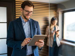 A man in a business suit and glasses is smiling while working on a tablet in a modern office. He is standing near a glass wall, with a woman in the background using her phone. The man appears focused and engaged, likely reviewing or analyzing something on the tablet. The setting is professional, with warm lighting and a sleek design, featuring wooden panels on the wall. The image conveys a sense of productivity, technology use, and a collaborative work environment.