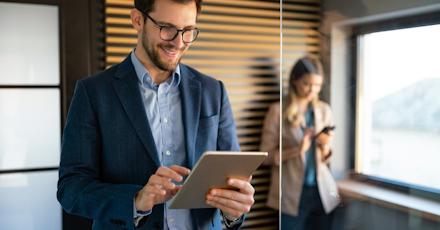 A man in a business suit and glasses is smiling while working on a tablet in a modern office. He is standing near a glass wall, with a woman in the background using her phone. The man appears focused and engaged, likely reviewing or analyzing something on the tablet. The setting is professional, with warm lighting and a sleek design, featuring wooden panels on the wall. The image conveys a sense of productivity, technology use, and a collaborative work environment.