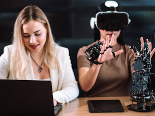 Woman using VR gloves and headset for robotics testing, while colleague works on laptop.