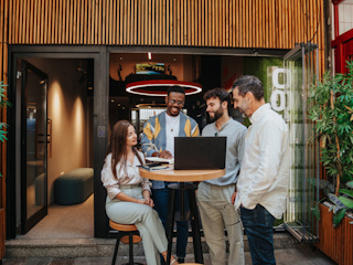 A diverse group of colleagues is gathered around a laptop during an outdoor meeting. The group consists of four men and one woman, all smiling and engaged in a collaborative discussion. They are standing and sitting around a high table in a modern outdoor space, with greenery and contemporary architecture in the background. The setting is casual and vibrant, reflecting a positive and creative work environment.