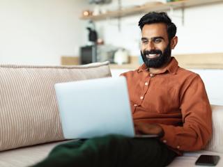 A man sitting on a couch smiles while shopping online using his laptop in a comfortable, modern home setting.