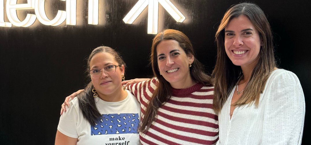 Three women team members smiling in front of a Valtech logo wall, highlighting leadership, diversity, and positive workplace culture.