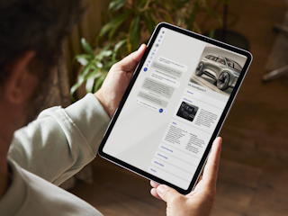 Person holding a tablet while browsing a car information app, displaying details and images of the VA GrandSpace X SUV.