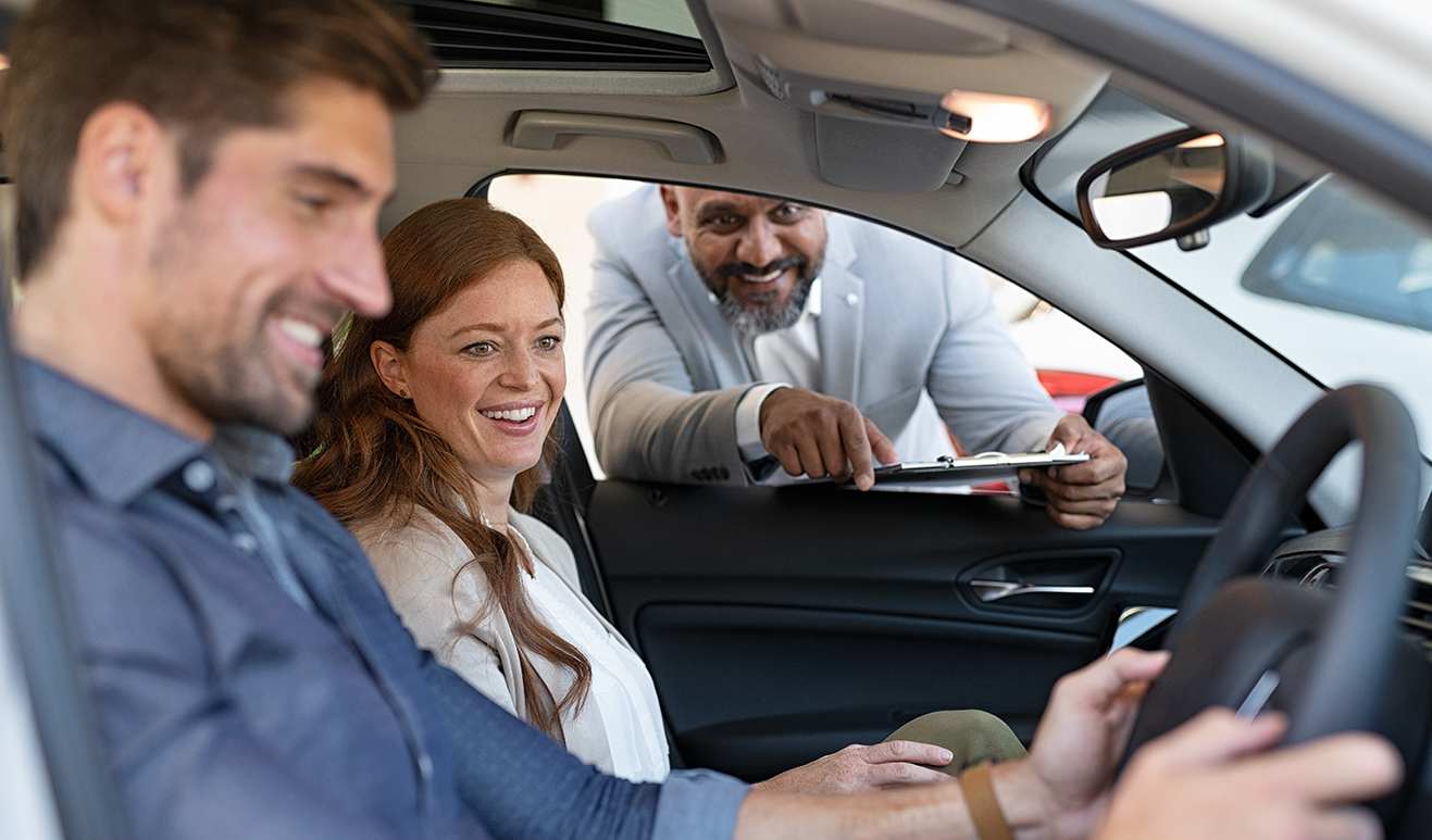 A man and a woman sit in the front seats of a European car with salesperson leaning in through the window pointing out features of the car