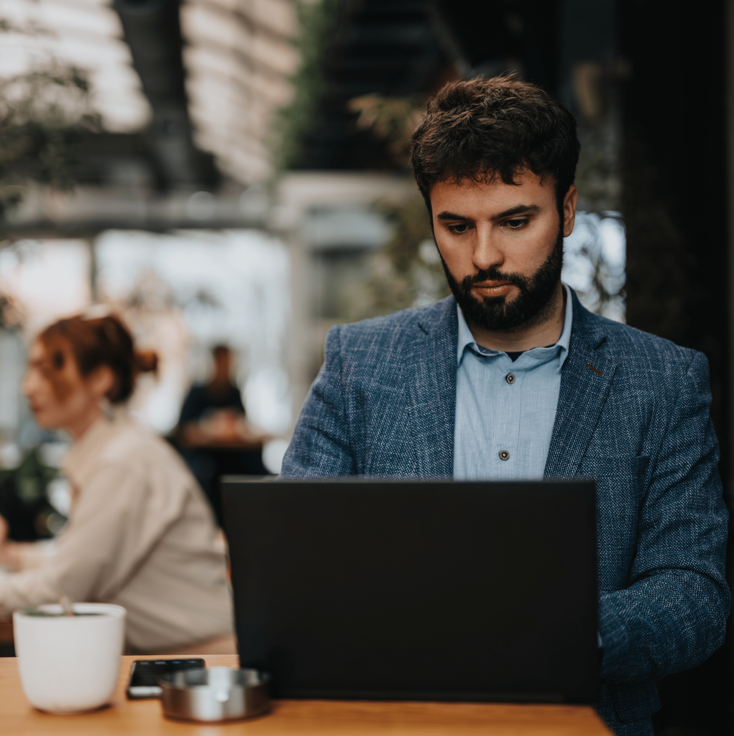  A man with a beard is focused on working on his laptop in a modern indoor setting, possibly a café or co-working space. He is dressed in a blue blazer and a light blue shirt, with a serious expression as he concentrates on his work. The background is blurred, showing other people and a warm, casual environment, with plants and soft lighting adding to the ambiance. A coffee cup and a smartphone are placed on the table next to him.