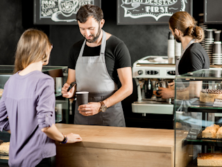 A barista in a gray apron preparing a coffee order for a customer at a modern coffee shop. The customer stands at the counter, while another barista works in the background near an espresso machine. The shop has a cozy, artisanal atmosphere with chalkboard menus in the background.