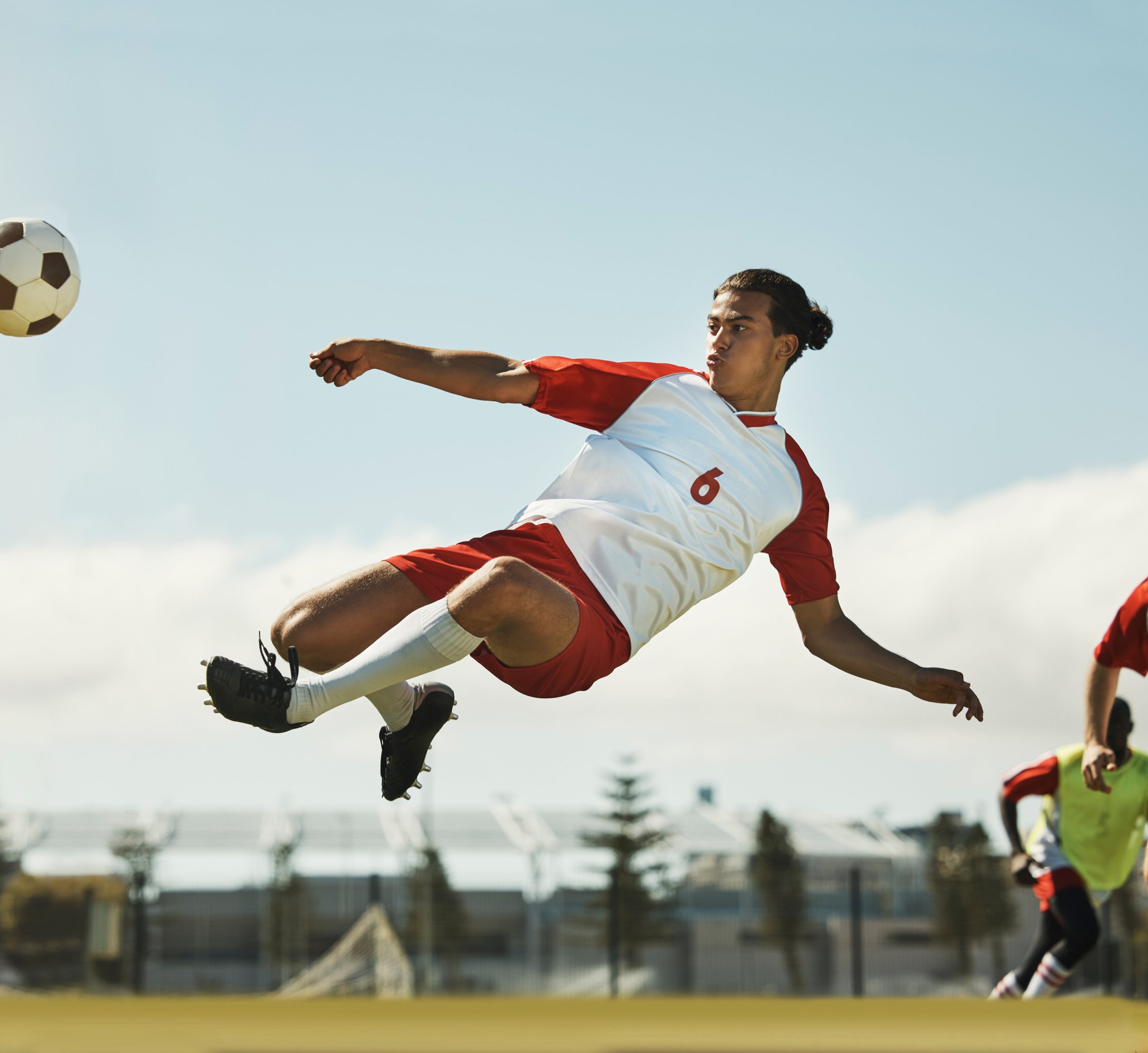 A soccer player in a red and white uniform is performing an acrobatic kick during a game. The player is airborne, fully extended, with intense focus on the ball as he makes contact. The background shows an outdoor soccer field with teammates and a clear, sunny sky, highlighting the dynamic action and athleticism in the moment.