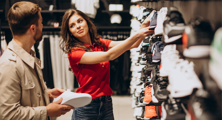 A woman in a red shirt helps a customer choose a pair of athletic shoes from a display in a retail shoe store.
