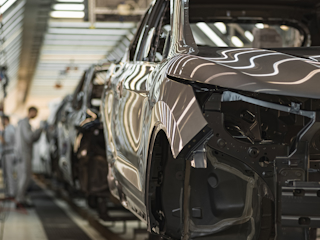 An automotive car assembly line in a factory with several vehicles in various stages of construction and workers blurred in the background.