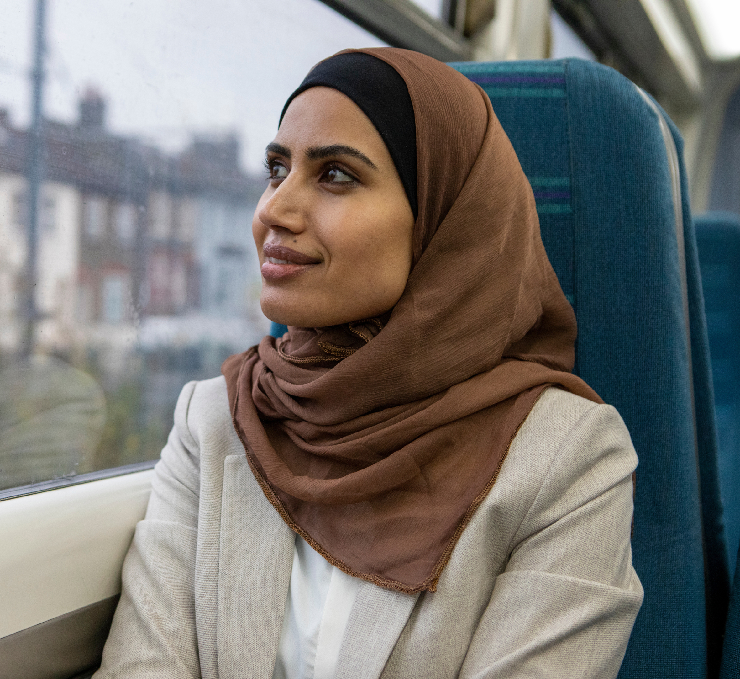 A woman wearing a brown hijab and a beige blazer sits on a train, looking out the window with a peaceful expression. The window shows a view of buildings blurred by rain, capturing a moment of quiet reflection during her journey.