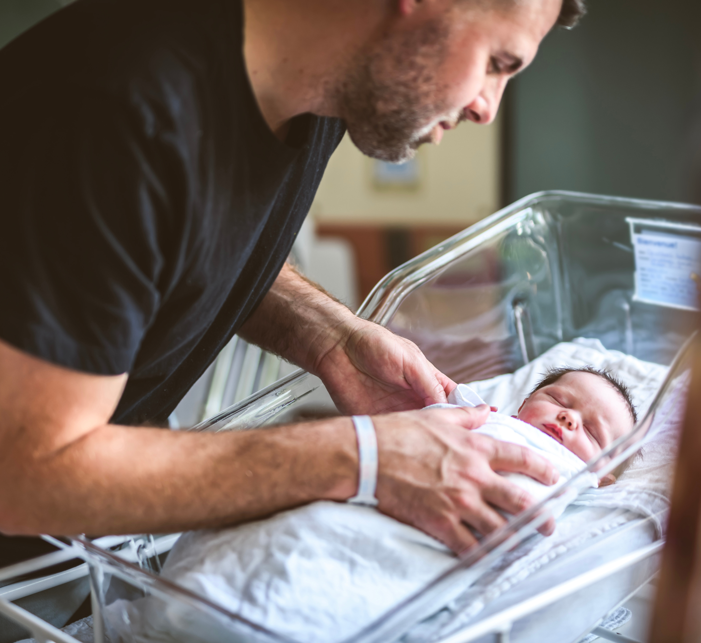 A father gently tends to his newborn baby, who is swaddled and resting in a hospital bassinet. The father is leaning over, carefully adjusting the baby’s blanket with tenderness and care. He is wearing a hospital bracelet on his wrist, indicating that this is likely soon after the baby's birth. The newborn is peacefully asleep, wrapped in a white blanket. The setting is a hospital room, with soft natural light creating a calm and intimate atmosphere. The image captures the bond between father and child in the early moments of life.