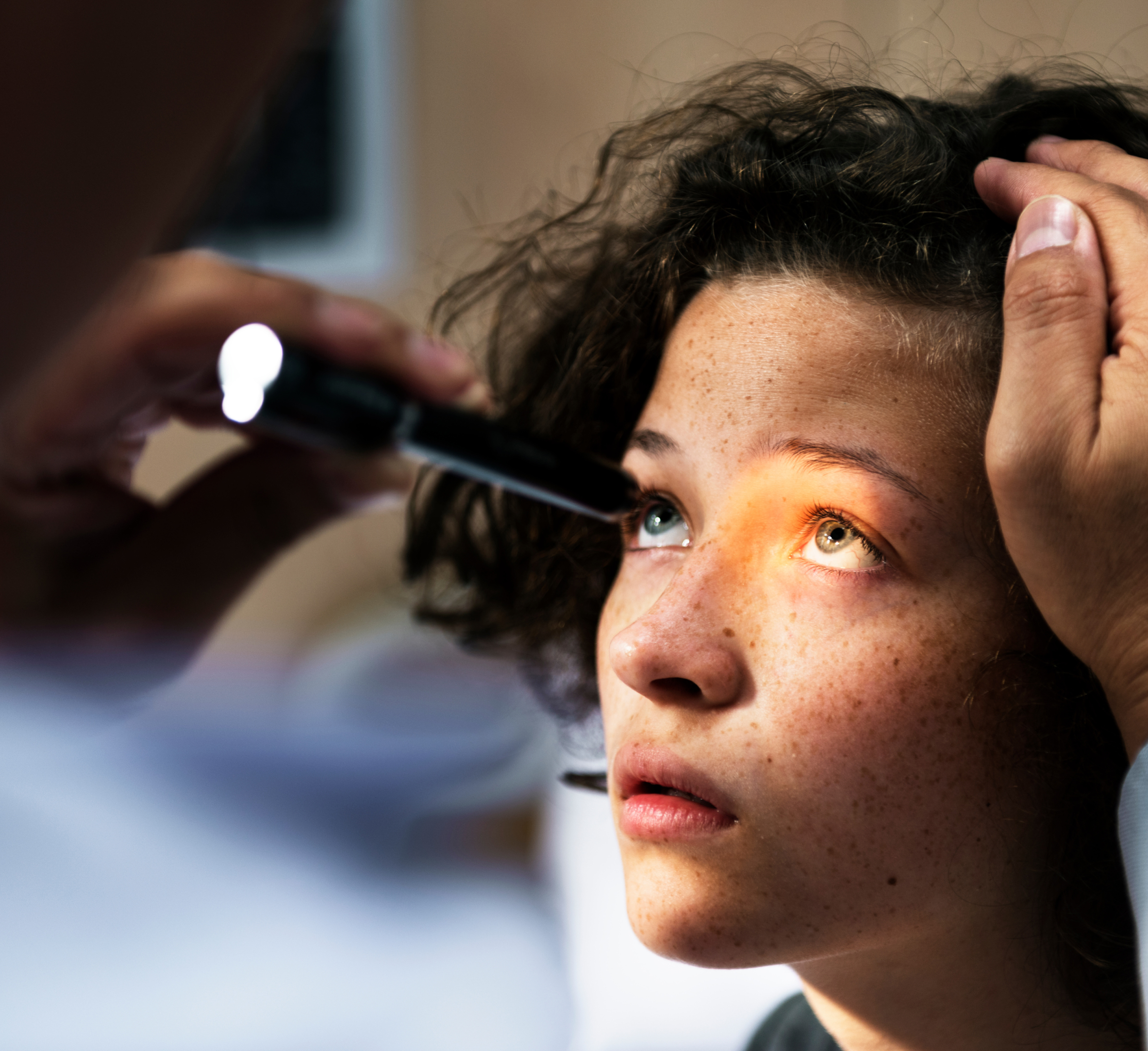 A young person is undergoing an eye examination. The doctor holds a flashlight close to the patient's eye, shining light to inspect the pupil's reaction. The patient looks upwards with wide, attentive eyes, while the doctor's hand gently holds their head steady. The setting is well-lit, and the image captures the detail of the eye inspection process, highlighting the patient's trust and the doctor's careful attention. The focus is on the importance of vision care and precise medical evaluation.