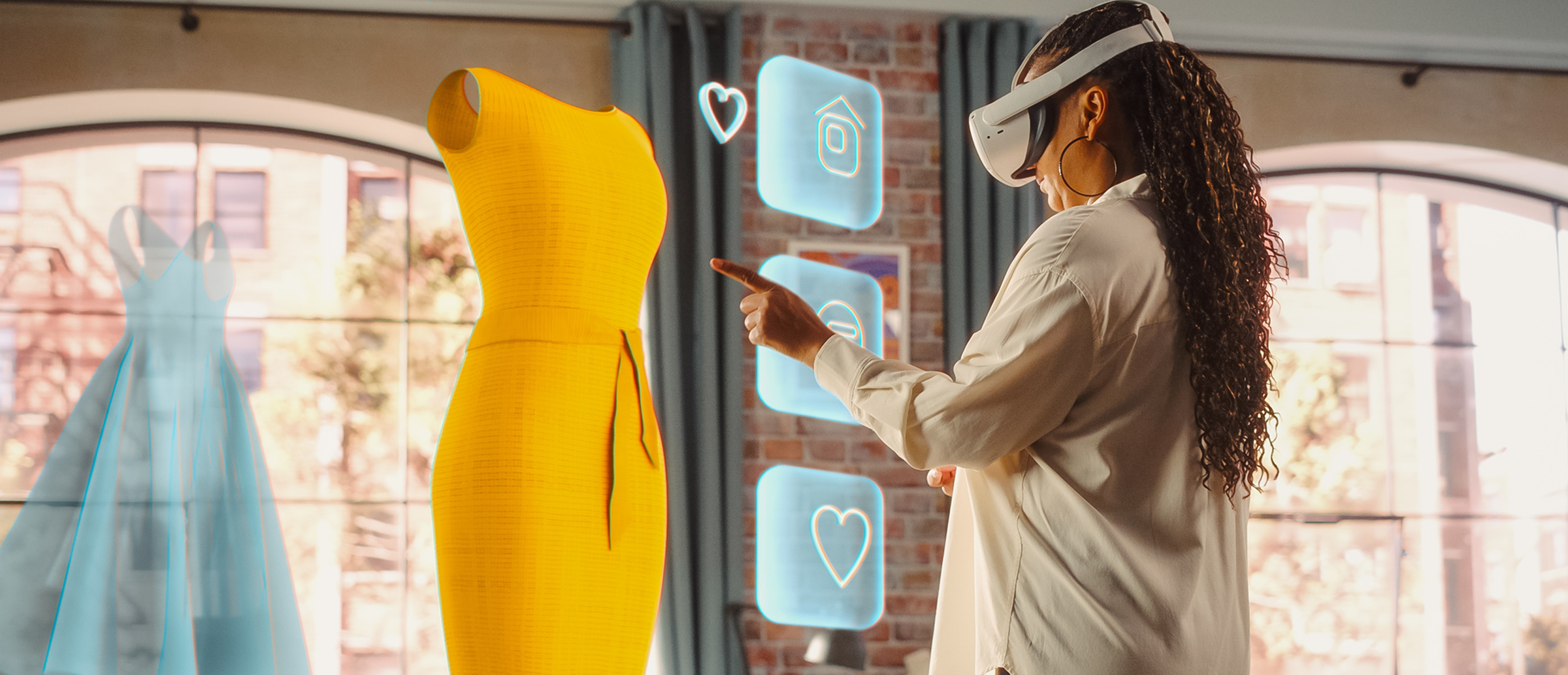 Woman wearing a virtual reality headset in a loft-style room, reaching out to navigate holographic dress models—one yellow and one blue—floating before large arched windows.