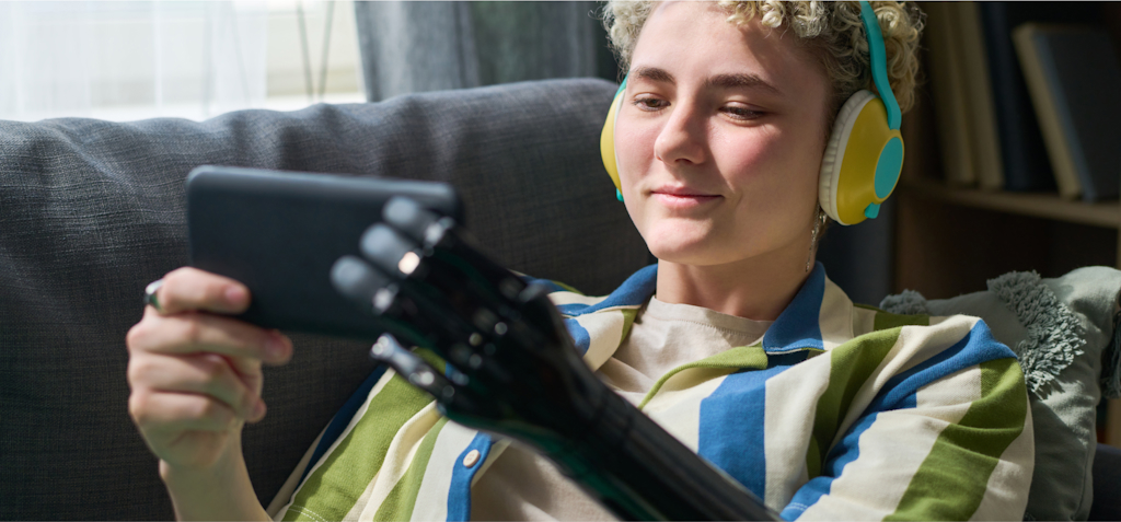 A young person with short, curly hair is lounging on a sofa, wearing bright yellow and blue headphones while watching something on their smartphone. They are smiling slightly, engaged in the content on their device. The person is holding the phone with a sleek black prosthetic hand, highlighting the use of assistive technology in everyday life. They are dressed casually in a striped shirt, and the setting is comfortable and relaxed, with soft lighting and a cozy atmosphere. The image emphasizes the seamless integration of technology in enhancing accessibility and entertainment.