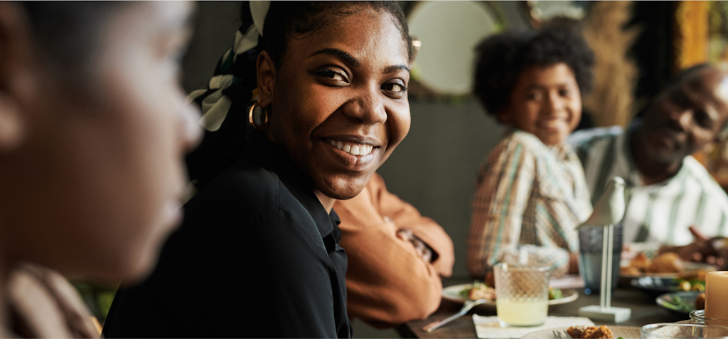 A young woman is smiling warmly at the camera while sitting at a dinner table with her family. She is wearing a black shirt and gold earrings, and her hair is tied back with a patterned scarf. In the background, other family members, including a young boy and an older man, are also smiling and enjoying the meal. The table is set with plates of food, drinks, and a candle, creating a cozy and welcoming atmosphere. The image captures the joy of shared moments and connection during a family gathering.