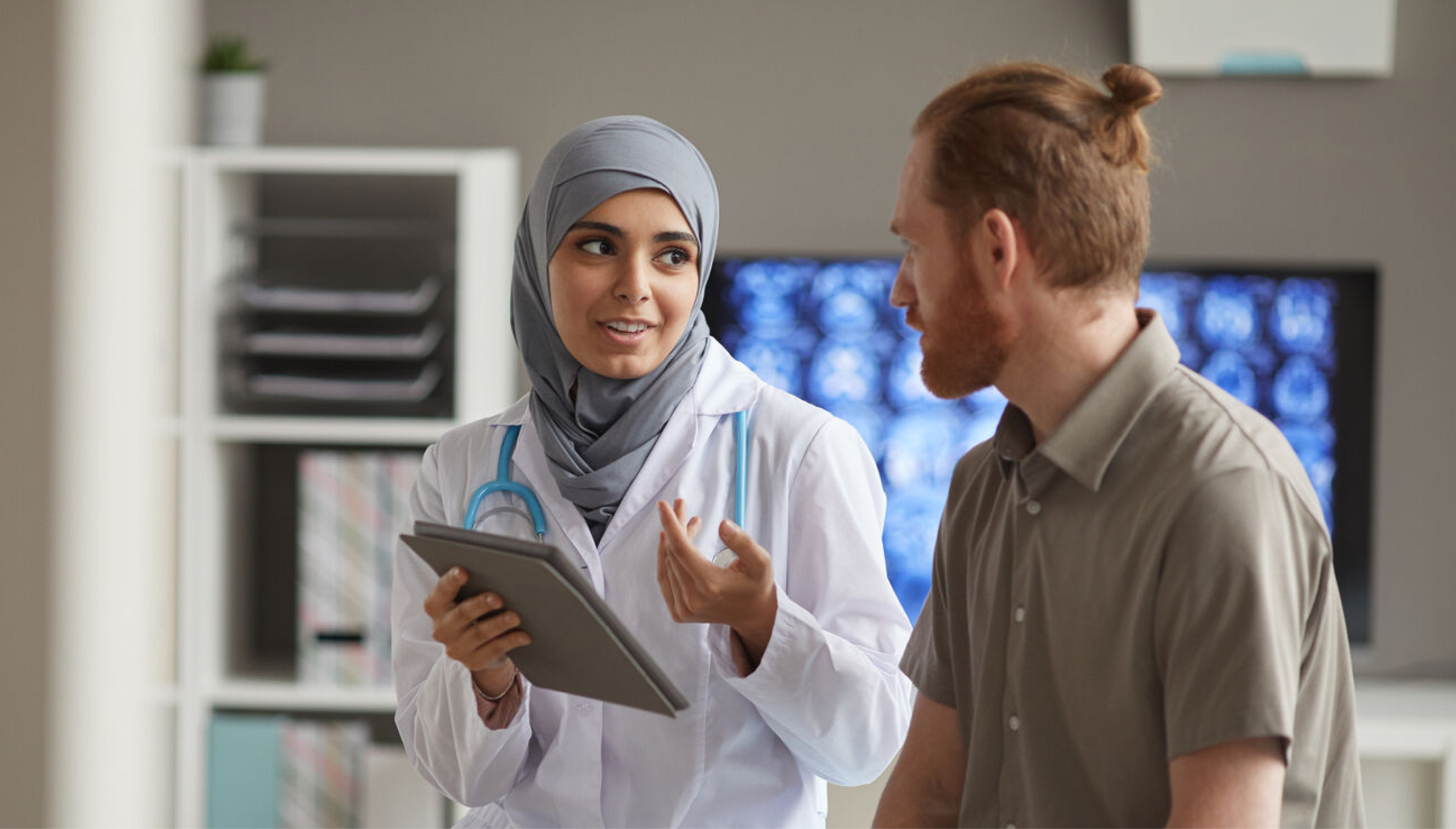 A female doctor wearing a hijab and a white coat with a stethoscope around her neck is engaging in a discussion with a male patient in a medical office. She is holding a tablet and explaining something with a friendly and professional expression. The patient, a bearded man with a tied-up hairstyle, is attentively listening. A screen in the background displays medical scans, indicating a diagnostic consultation.