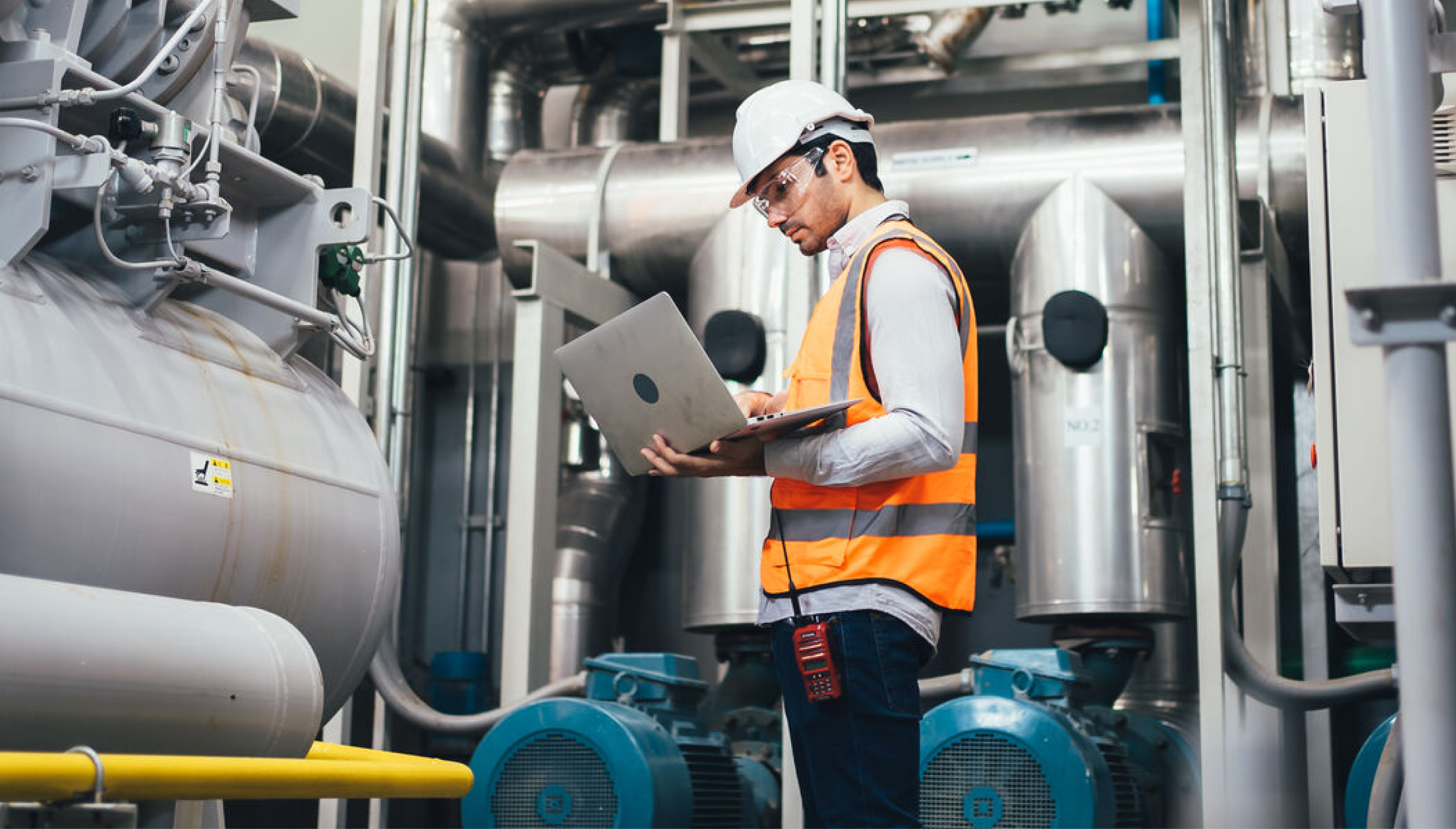 A male industrial engineer wearing a white hard hat, safety glasses, and an orange reflective vest is working on a laptop in a factory or industrial facility. He is surrounded by large machinery, pipes, and ventilation systems. A walkie-talkie is clipped to his waist, indicating a role that requires real-time communication. The setting suggests an inspection, maintenance check, or system monitoring in a manufacturing or energy plant.