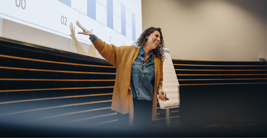 A woman is standing in a lecture hall or conference room, delivering a presentation.
