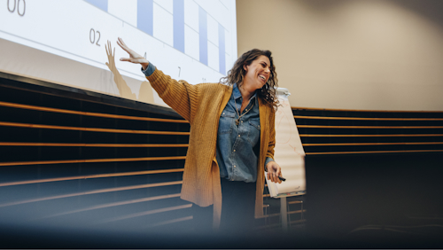 A woman is standing in a lecture hall or conference room, delivering a presentation.