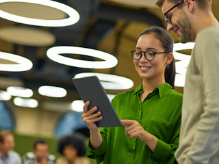 A smiling woman in a green blouse holds a tablet while standing next to a man in glasses and a light sweater, both looking at the screen together in a modern office space with circular overhead lights and a blurred group working in the background.